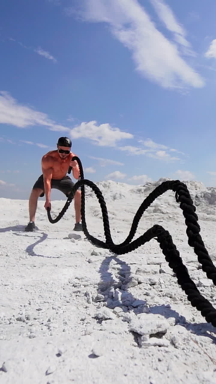 Muscular man with black ropes on white hill under blue sky. Athletic male performs Battle Ropes exercise during an outdoor cross fitness workout. Slow motion. Vertical video