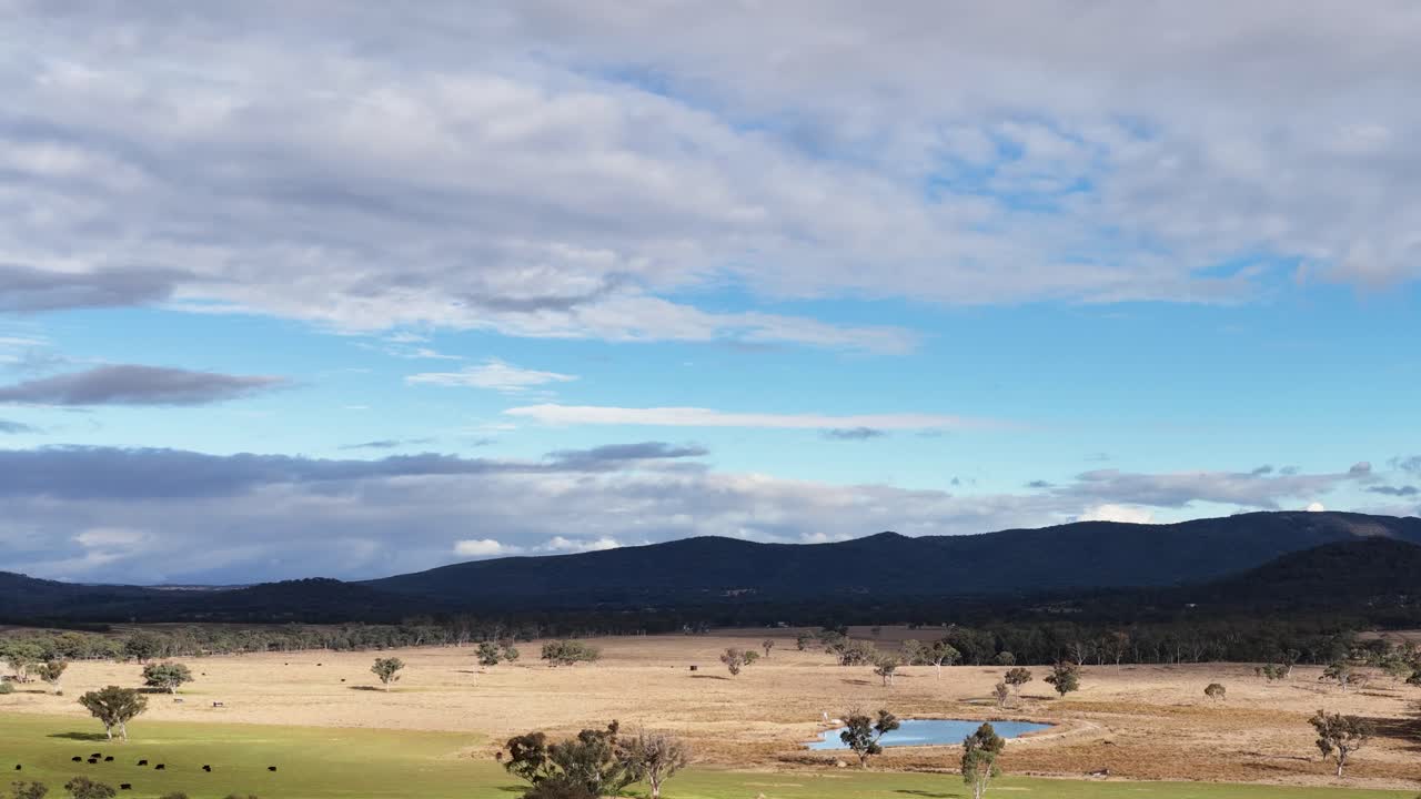 Wide shot of people engaged in clay target shooting near a farm pond, surrounded by open fields and distant hills under partly cloudy daylight