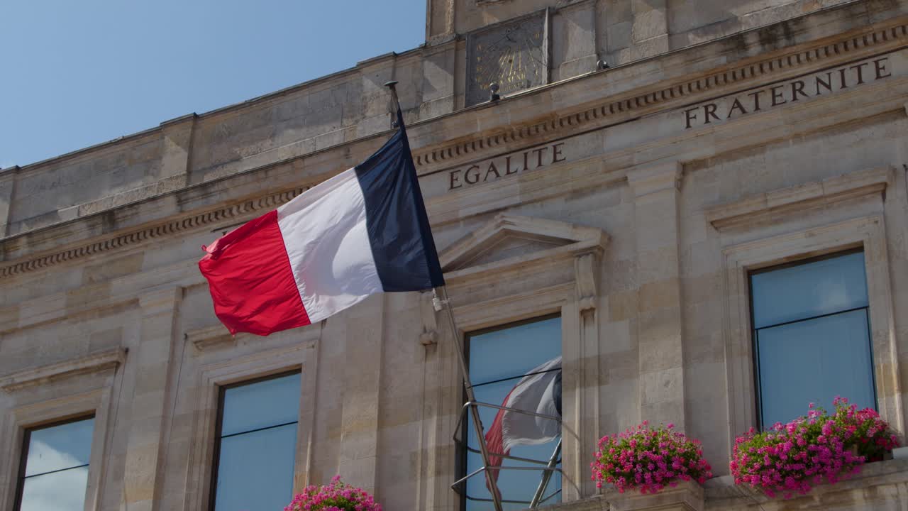 French tricolor flag waves outside classic stone town hall, bright daylight, steady camera, floral decorations