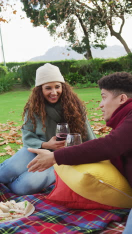 video vertical de una feliz pareja biracial haciendo picnic, bebiendo vino en el jardín de otoño
