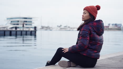 an asian woman relaxes by the waters of valencia port, spain, wearing a red beanie that complements her matching checkered jacket, casting a serene gaze towards the camera