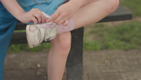 girl sitting on bench wearing pink shoe over sock, close up leg view, mother assisting, summer park setting, focus on foot and strap adjustment, casual moment of dressing before play