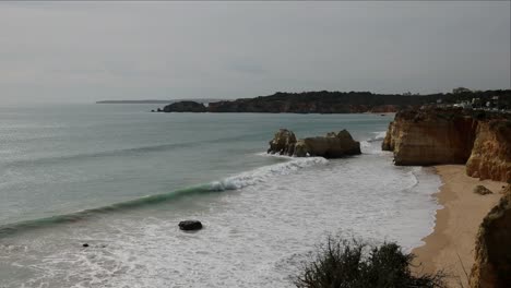 Amplia-Vista-En-Cámara-Lenta-Desde-Los-Acantilados-Sobre-Praia-Do-Vale-Do-Olival,-Portugal-En-Una-Tarde-Nublada-Con-Horizonte-Nublado-Sobre-El-Agua-Y-Olas-Rompiendo-Contra-Enormes-Rocas