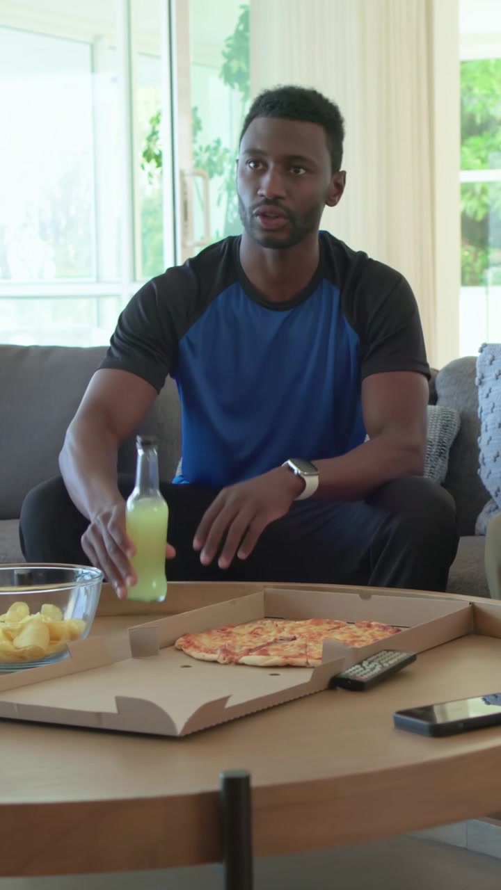 Vertical video: Bringing green drinks man in blue opening pizza box home, sitting and sharing chips