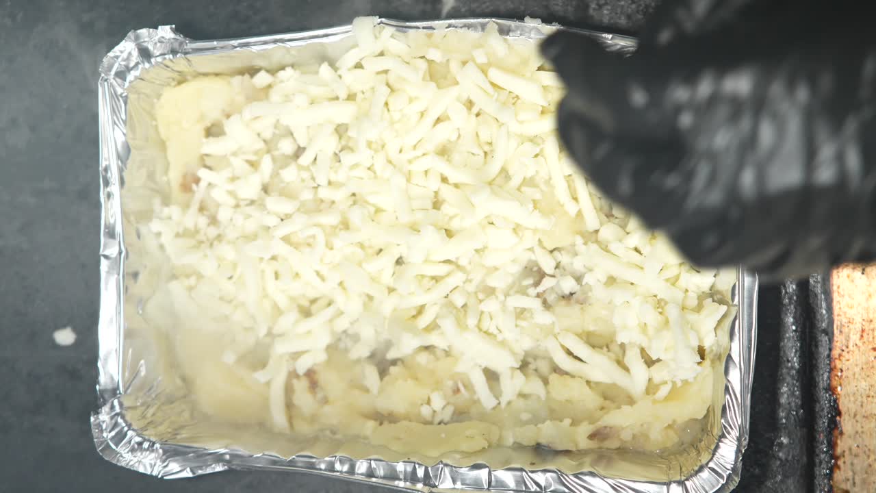 Manos con guantes higiénicos añadiendo queso rallado a una papa loca. Este snack de comida callejera mexicana incluye patata asada, mantequilla, crema agria y salsa, resaltando ingredientes frescos y sabores vibrantes.