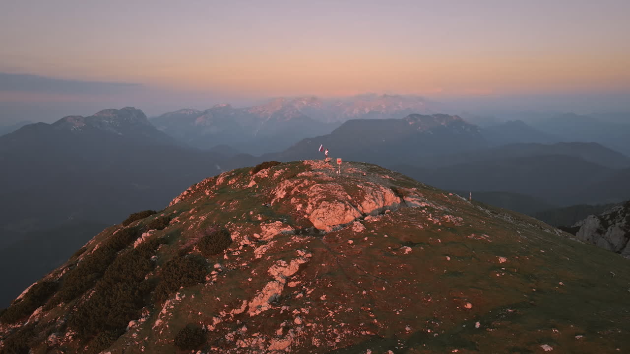 toma de drone de la cima de la montaña peca en el sunrse donde un excursionista sostiene una bandera eslovena en el poste