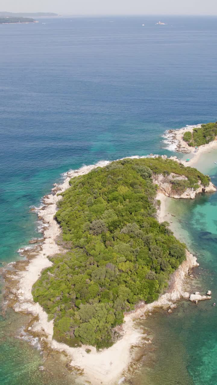 Vibrant green Ksamil island surrounded by crystal-clear turquoise waters under a sunny sky. Aerial, Vertical Video