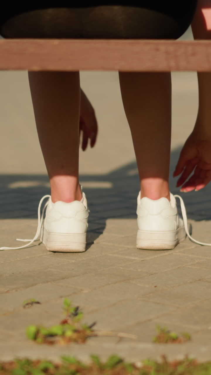 밝은 빛 아래 벤치에 앉아있는 하 스니커의 신발을 풀고있는 여자의 다리 뒷면, 보행로에 부드러운 그림자가 여 있고 롤러 스케이트는 벤치에서 부분적으로 볼 수 있습니다.