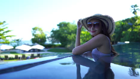 atractiva mujer asiática con un sombrero a rayas y grandes gafas de sol sonriendo mientras disfruta de una piscina infinita a la sombra con agua cristalina y luz solar brillante en los árboles verdes en el fondo