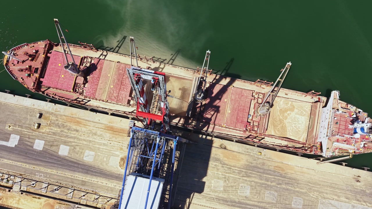 Large cargo ship is being loaded at a dock in a port with cranes and equipment in bright daylight