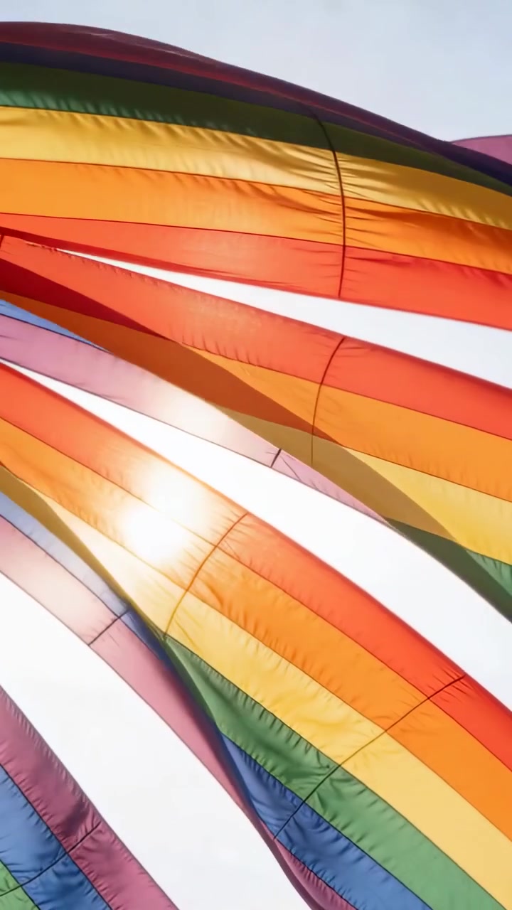 Vertical video: Billowing rainbow-flag fabric shifting in wind over open sky, showing sun glare