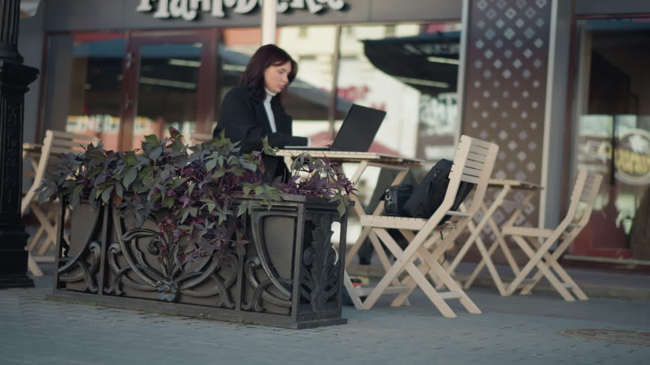 estudiante con cabello oscuro escribiendo en una computadora portátil en una mesa al aire libre en un ambiente acogedor de centro comercial, rodeado de plantas y sillas con un fondo borroso con una inscripción visible en la pared