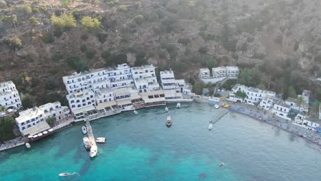 Drone-view-in-Greece-flying-over-blue-sea-in-Loutro-small-white-house-town-and-small-boats-next-to-a-hill-on-a-sunny-day