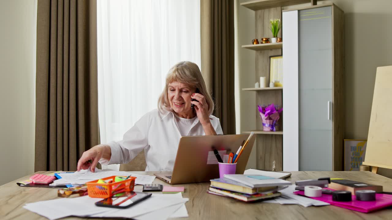 Elderly businesswoman working from home online and using her smartphone as she does paperwork during the lockdown for Covid-19 or coronavirus during the pandemic