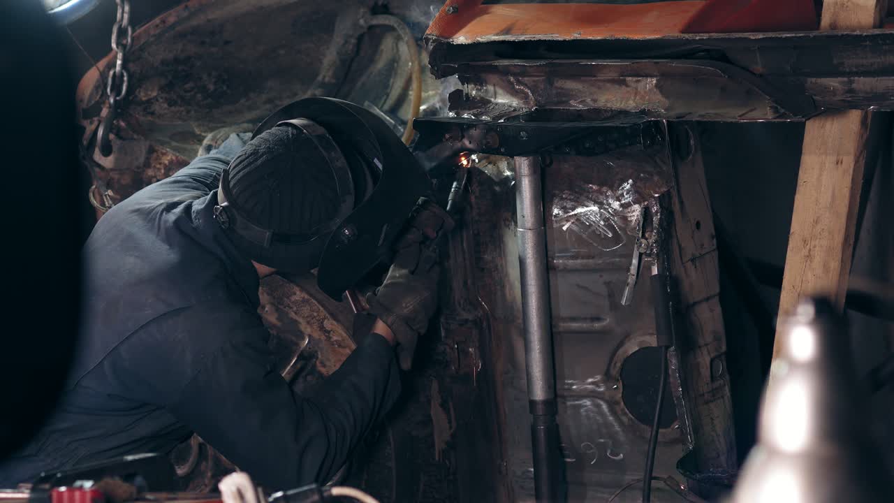 Backside footage of a welder joining some metal pieces together on car's bottom surface with a welding torch.