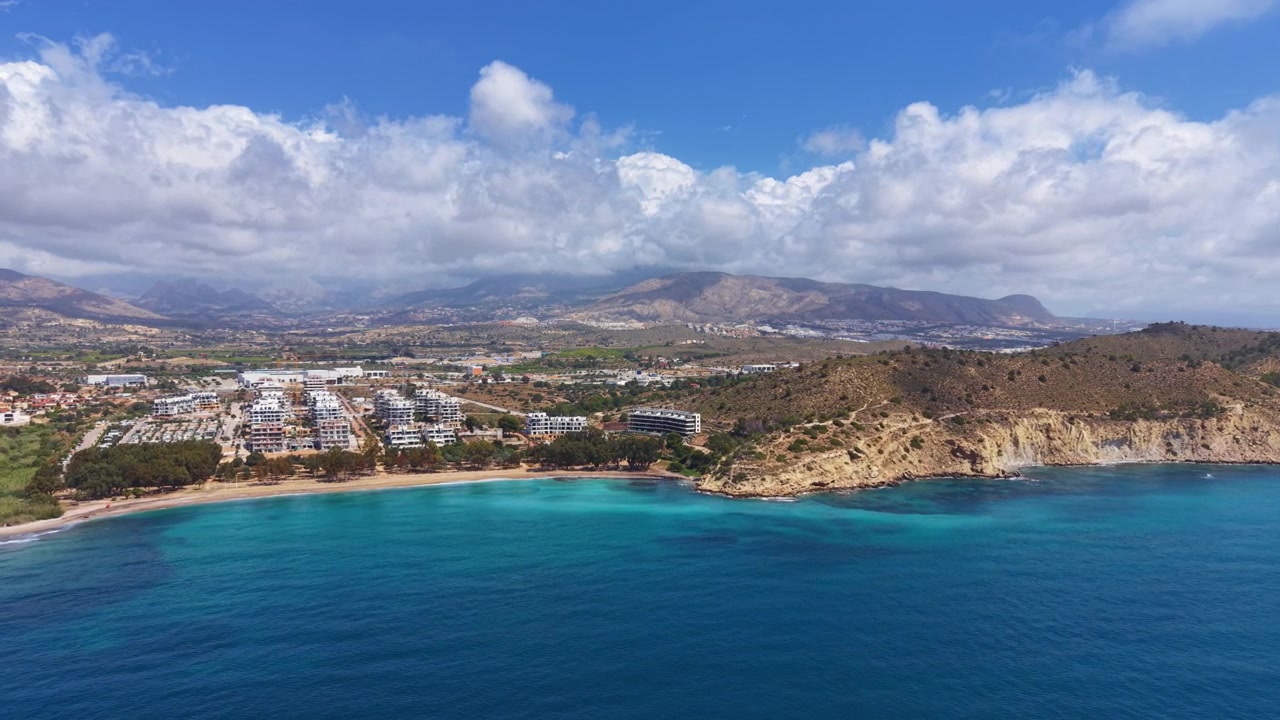 Baía mediterrânea turquesa ladeada por penhascos rochosos e edifícios residenciais costeiros modernos, com montanhas e nuvens brancas estendendo-se pelo horizonte