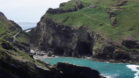 las ruinas del popular destino turístico castillo de tintagel en cornwall en la cima de un acantilado con un nuevo puente que conduce a la isla y la bahía con playa debajo