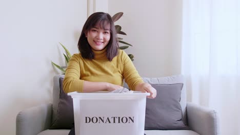 portrait of asian young woman looking at camera holding box with clothes for donation