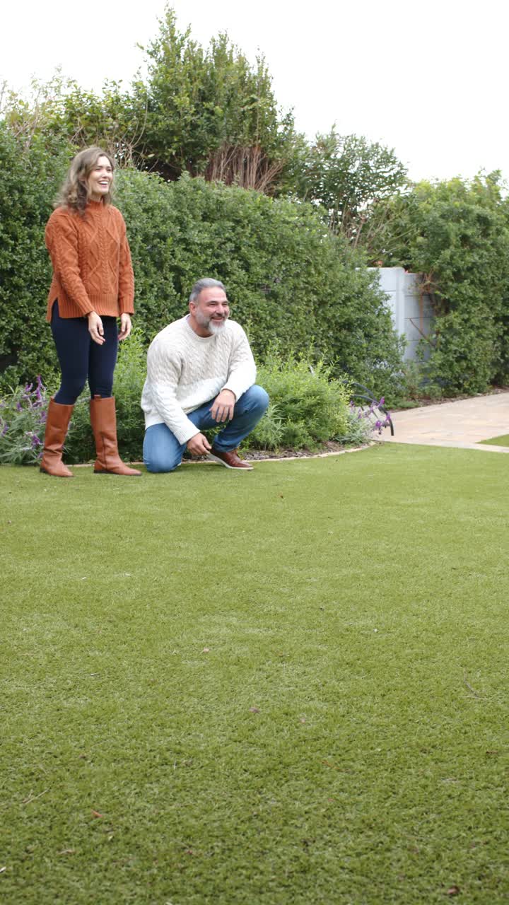 Vertical video: Couple kneeling on lawn offering ball for dog to fetch, tossing it, copy space
