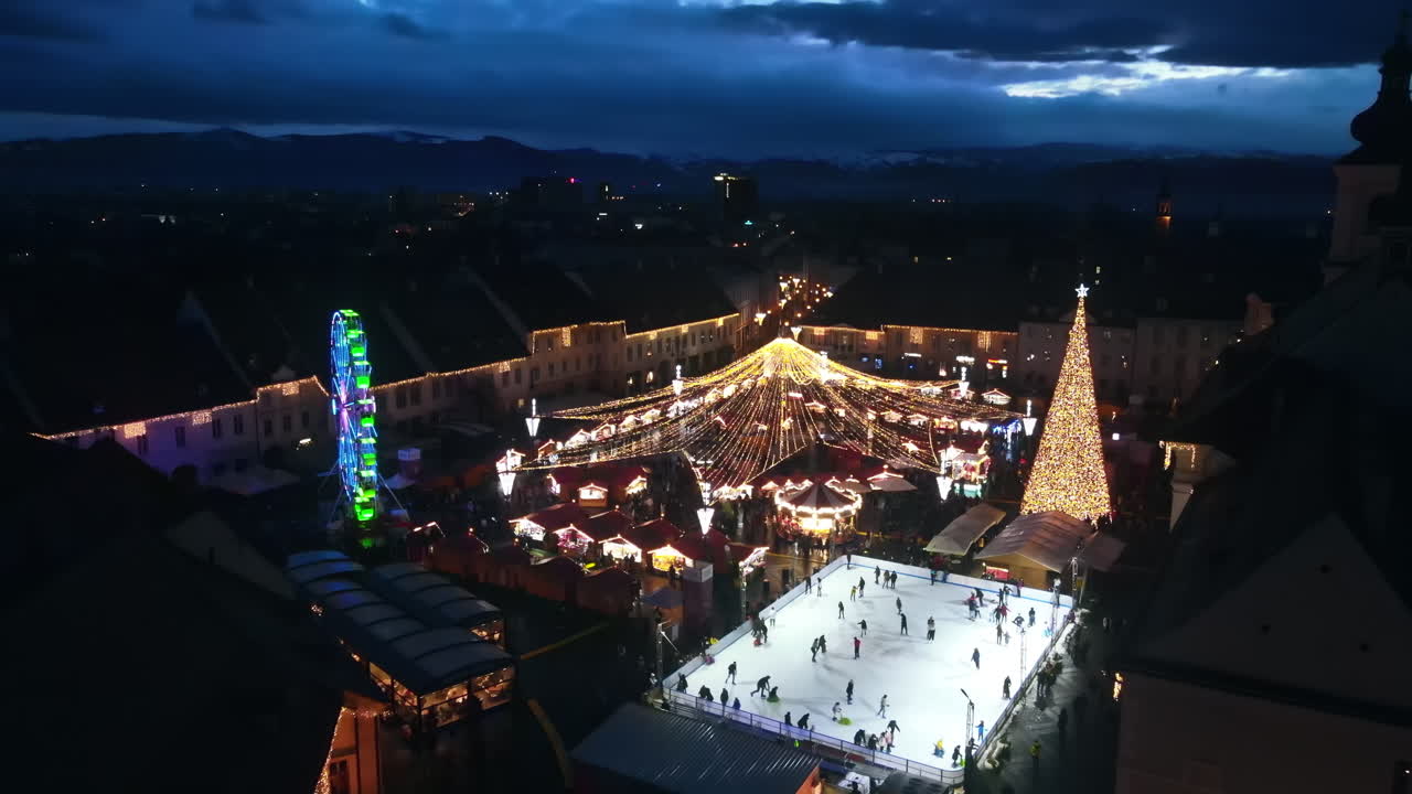Aerial drone view of The Big Square in Sibiu at night, Romania. Old city centre decorated for Christmas. Ferris wheel, skating rink, people