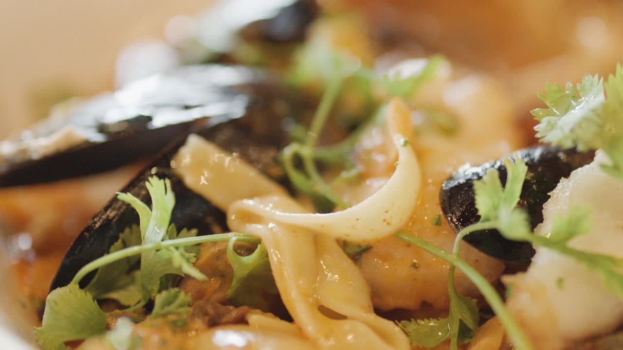 Extreme close-up of assorted seafood including mussels, squid, and scallops in a steaming chowder, garnished with fresh herbs under warm, soft lighting