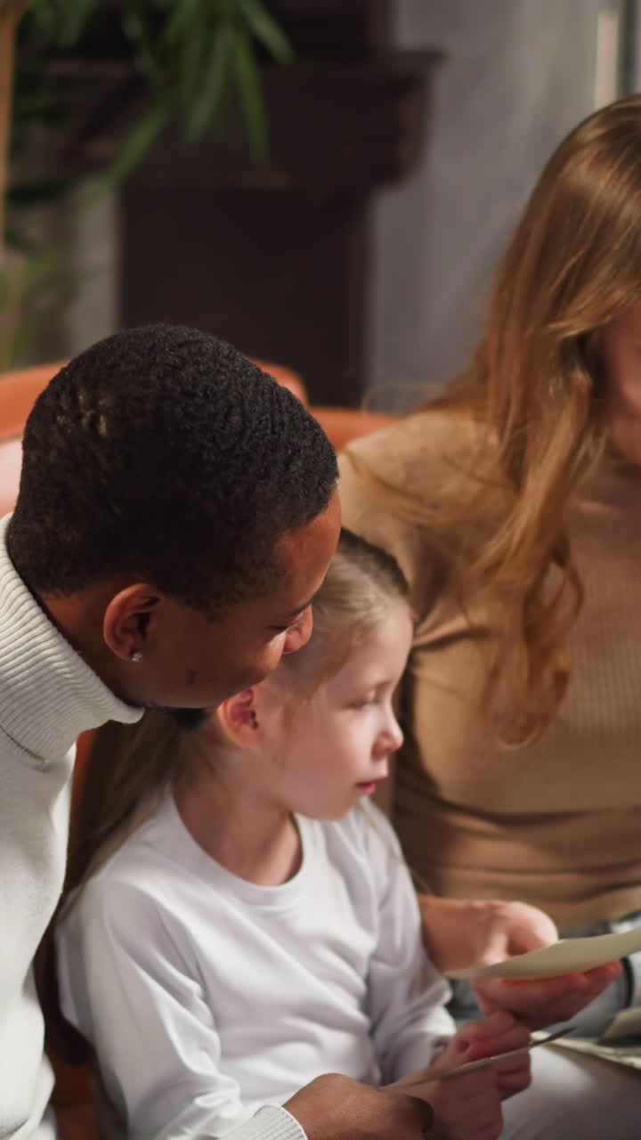 Preschooler girl shows relatives on old photos enjoying family memories with African-American father and white mother at home slow motion