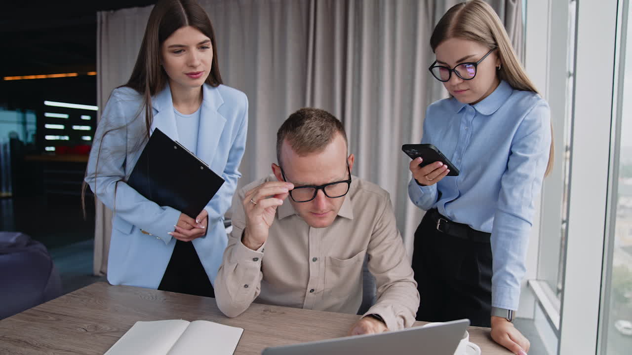 Male manager looking at laptop in surprise, putting off his glasses. Ladies colleagues standing beside him. Cooperation in the office and teamwork.