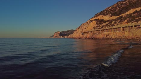 Amanecer-Dorado-En-La-Playa-De-Port-Ginesta,-Barcelona,-Con-Olas-Tranquilas-Y-Acantilados-Rocosos.