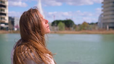 Young-beautiful-woman-enjoys-sunshine-near-city-water-pond,-close-up-slow-motion-view