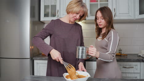 young girl helps her aunt in kitchen, holding metal sifter while aunt stirs batter in bowl, they work together in bright, modern kitchen, showcasing family bonding