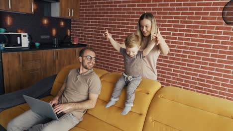 mother plays with her son distracting their father busy with laptop on the sofa