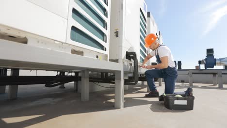el joven ingeniero está manteniendo el sistema de aire acondicionado. está girando el tornillo con una llave. está ubicado en el techo del centro de negocios. hay una caja de herramientas al lado. lleva ropa de trabajo y un sombrero. buen día soleado. 4k