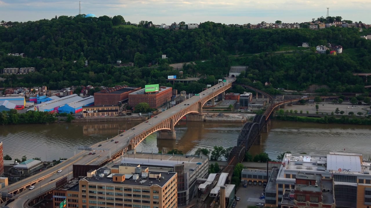Pittsburgh, Pensilvanya'da bir şehir üst geçit köprüsü, nehir kenarı binaları ve yemyeşil tepelere yukarıdan bakan yüksek havadan drone görüntüsü.