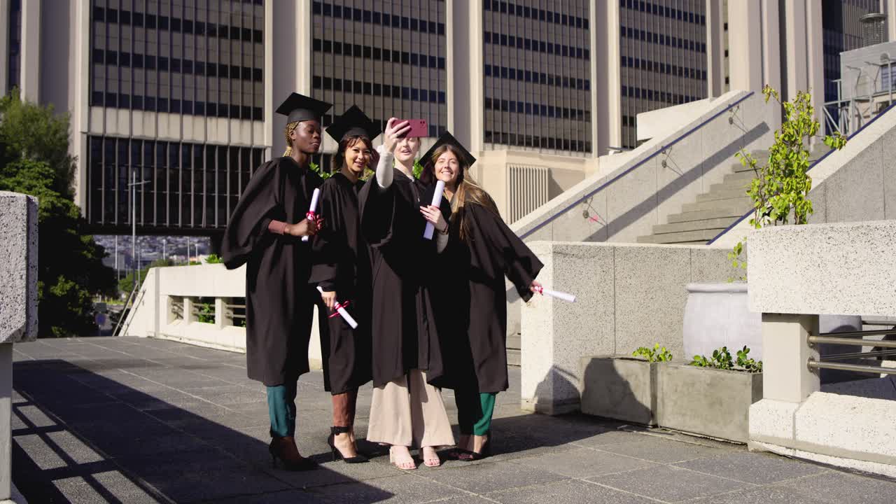 Diverse women in gowns crowding peer phone on plaza posing for selfie, raising diplomas for grad