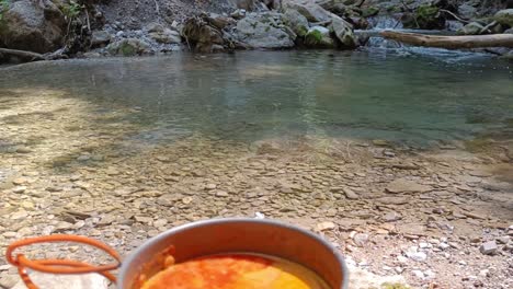 primer plano de la cocina hobo cocinando sopa de tomate en una olla de cocina de camping chino con cascada y río en segundo plano.