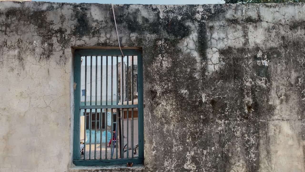 A shot of house in a village. Old, textured wall with a window and a woman is seen sitting inside the yard