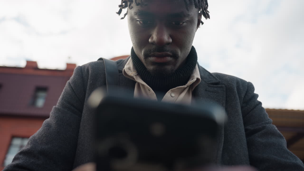 Sudden blurry view of male figure holding smartphone outdoors, wearing dark coat and shirt under daylight, conveying motion, focus, and mood of digital engagement with natural light ambiance