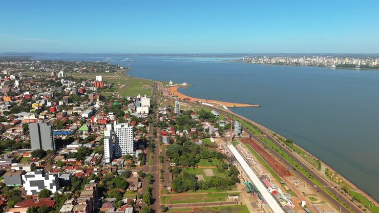 Encarnación, Itapúa, Paraguay, näyttäen kaupunkikuvan, ranta-alueen ja rannan Paraná-joen varrella, Posadasiin, Argentiinaan johtavan sillan näkyessä etäisyydessä kirkkaan taivaan alla, ilmakuvaus liukuen.