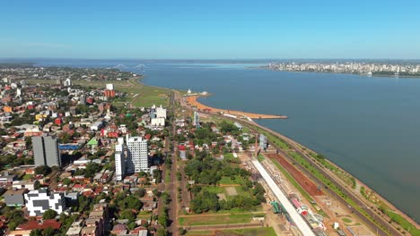 encarnación, itapúa, paraguay, showing urban skyline, beach area, and waterfront along the paraná river, with the bridge to posadas, argentina, visible in the distance under a clear sky, aerial glide