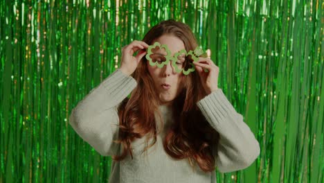 woman celebrating st patrick's day standing in front of green tinsel curtain wearing prop shamrock shaped glasses 1