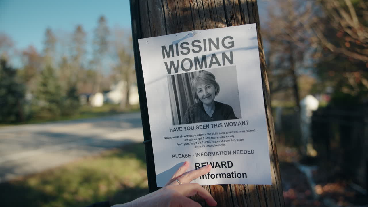 Mãos removendo um cartaz de mulher desaparecida de um poste de madeira em uma rua suburbana, sugerindo um desaparecimento ou uma atualização na investigação.