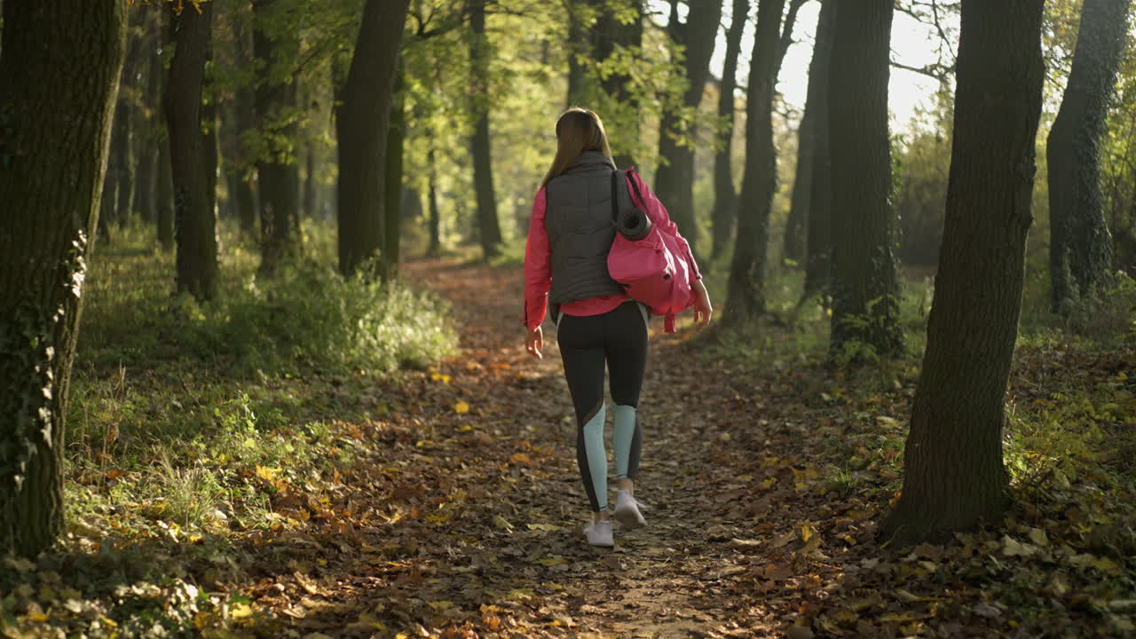 Woman walks on a path in an autumn forest carrying a yoga mat