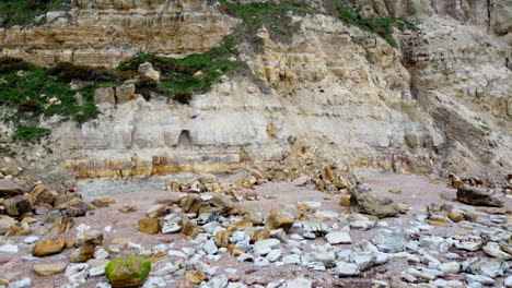 una vista aérea se eleva desde una playa rocosa y sube por un acantilado con estratos visibles, afloramientos de hierba y desprendimientos de rocas para revelar un acantilado coronado con hierba verde, arbustos y árboles