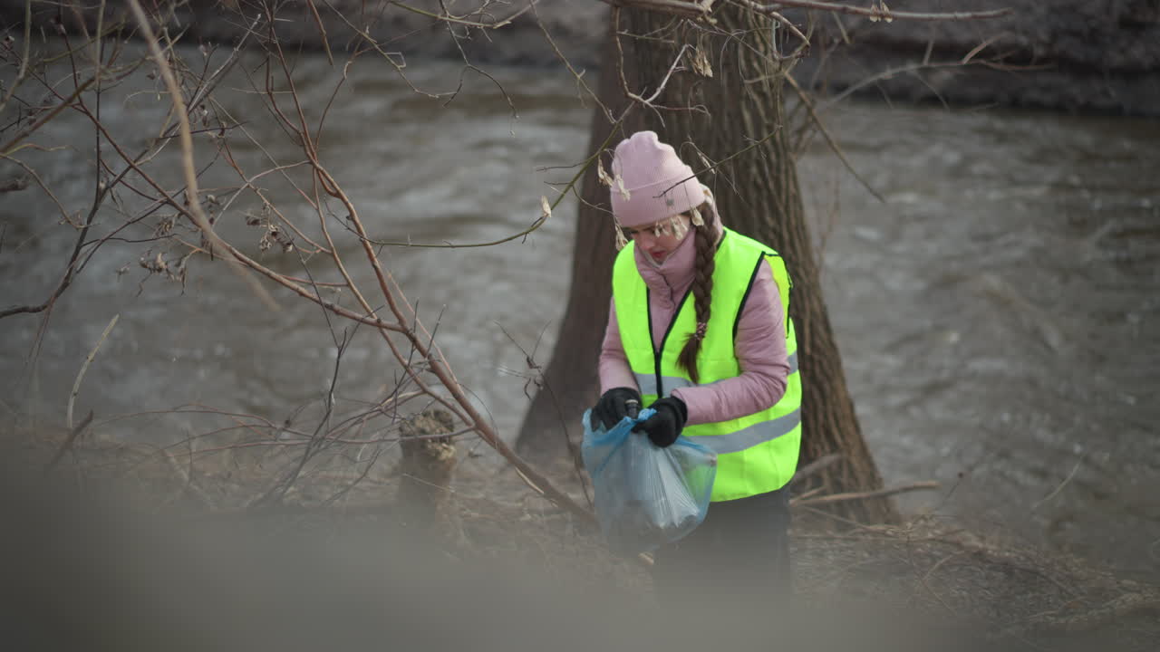 Woman wearing neon safety vest and pink beanie collects trash in blue plastic bag on riverbank during environmental cleanup, bending down among branches and debris to remove litter