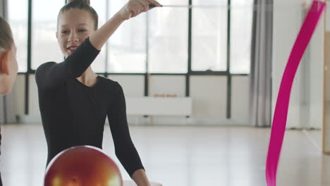 Gymnastic-Blonde-Girl-Playing-With-A-Band-While-Talking-With-Her-Friend-Before-Starting-Ballet-Class