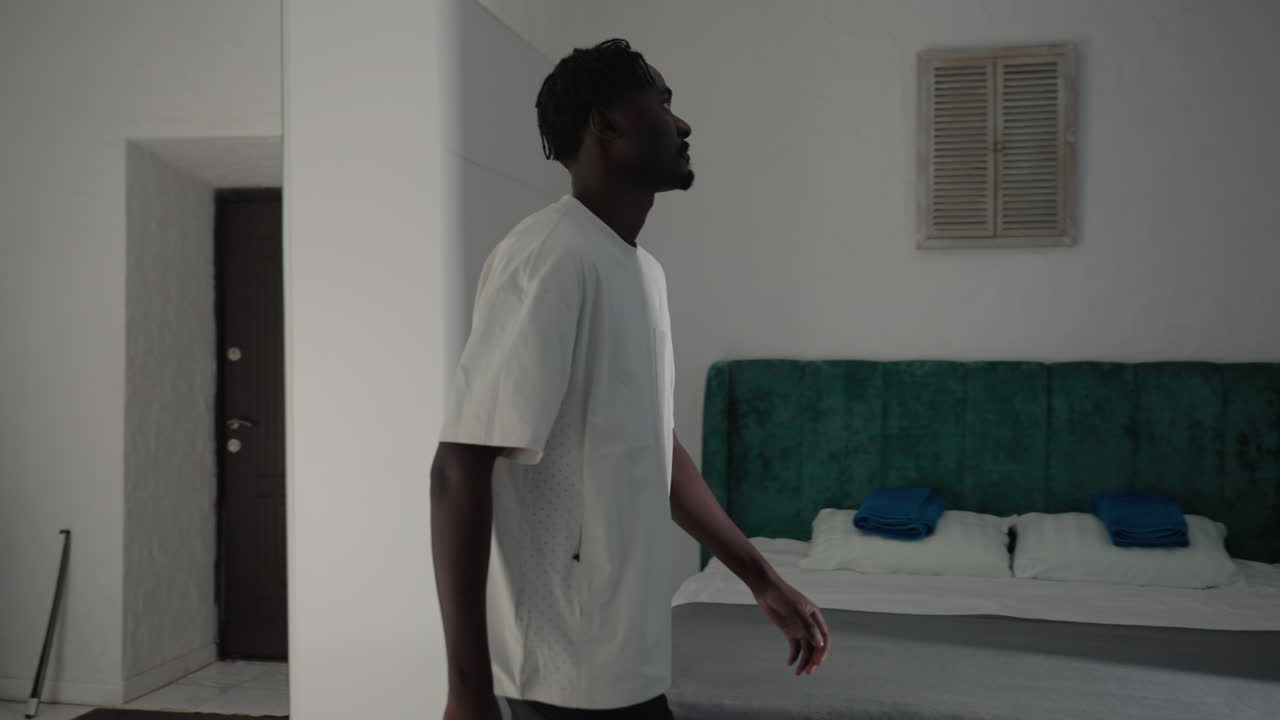 Man in white t-shirt and black shorts bending down to grab his black backpack in minimalist kitchen, preparing to leave home, sunlight streaming in, simple furnishings and doorway visible