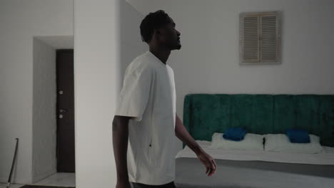 man in white t-shirt and black shorts bending down to grab his black backpack in minimalist kitchen, preparing to leave home, sunlight streaming in, simple furnishings and doorway visible