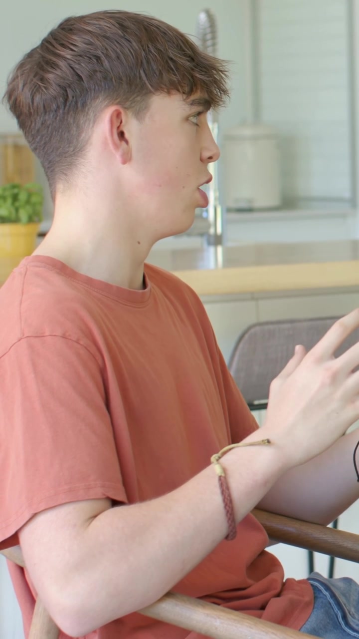 Vertical video: Hearing voice offscreen, boy orange tee replying on chair at counter bracelet and pl