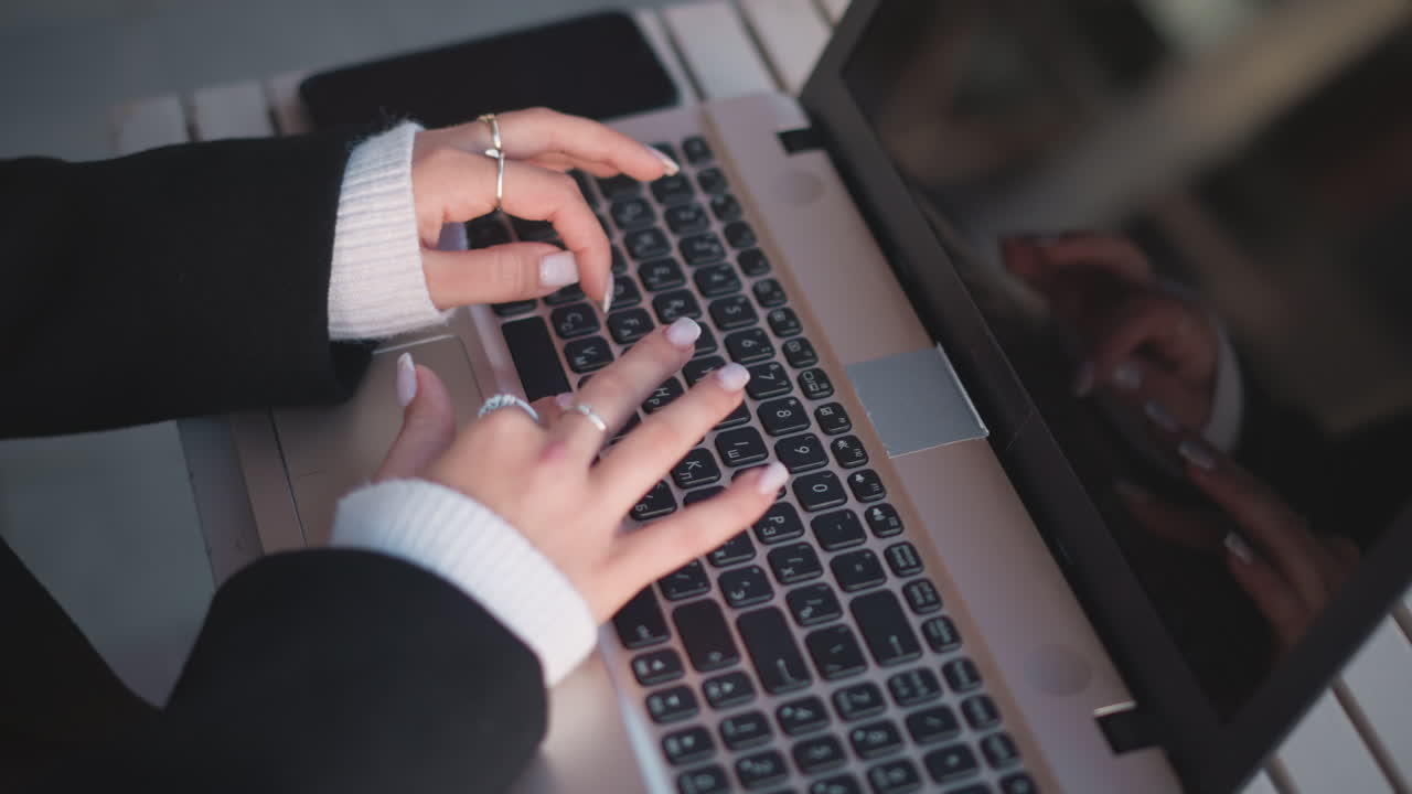 섬세한 반지와 은 손으로 장식된 손의 클로즈업, 노트북 키보드에서 타이핑, 화면에 반사, 쾌적한 스웨터 팔꿈치가 따뜻함을 더하고, 나무 테이블에 스마트폰이 근처에 있습니다.