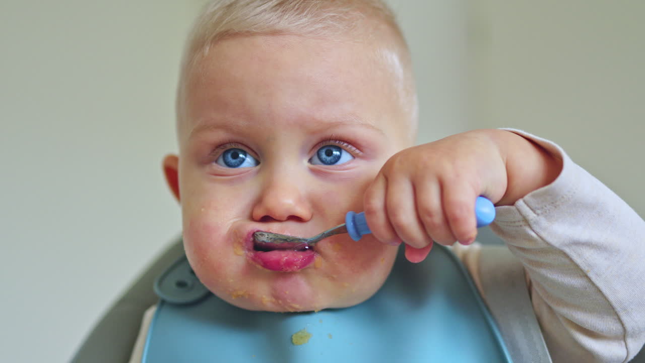 Cute one-year-old boy sits in a high chair and eats independently. Silicone baby feeding utensils. Child learns to eat independently